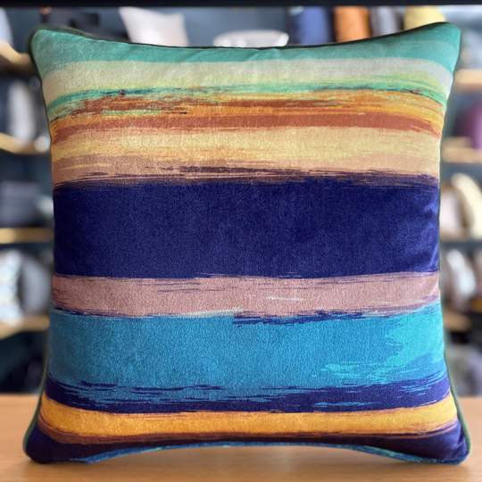 A square-shaped throw pillow with a geometric pattern featuring bands of blue, orange, and green velvet, with piping edges on the corners.